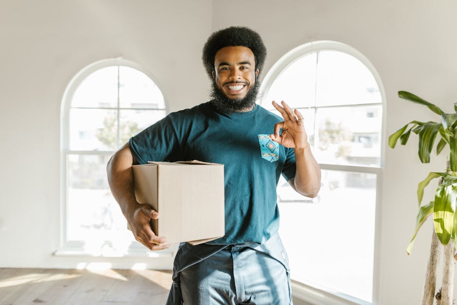 A smiling man with a beard and curly hair standing inside a bright room with large arched windows, holding a small cardboard box. He is wearing a dark teal t-shirt and grey trousers, and making an 'OK' gesture with his right hand. Various green indoor plants are visible on the right side of the image. The room has a light-colored wooden floor and white walls, with sunlight streaming through the windows. The scene depicts a home relocation or furniture transport process, with the man representing a professional moving service, such as that provided by Man and a Van Haringey, during packing and loading activities.