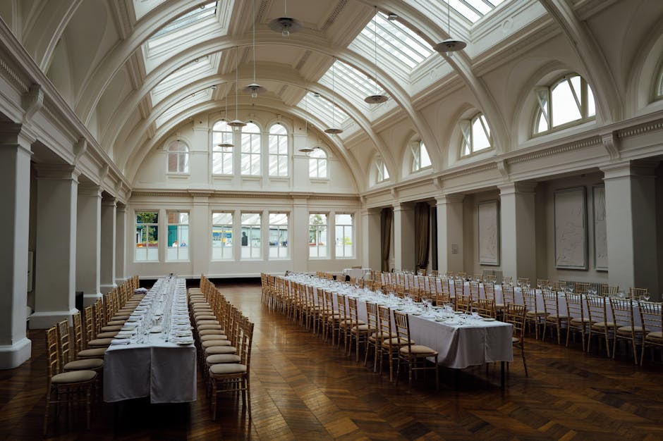 A spacious, elegantly decorated interior with a high, arched ceiling featuring large, natural light-filled windows and intricate architectural moldings. The room contains multiple long, rectangular tables arranged in parallel, covered with white tablecloths and neatly set with glassware, plates, and cutlery for a formal event. Surrounding the tables are gold-colored chairs with padded seats, aligned to prepare for a seated gathering or banquet. The polished wooden floor adds warmth to the space. Outside the windows, a glimpse of the street and greenery can be seen, indicating this is a venue suitable for house removals and event relocations. The interior is well-lit by natural daylight, highlighting the detailed ceiling structure and spacious layout. Man and a Van Haringey often assists with venue moves, including sensitive packing, furniture transport, and loading logistics, ensuring seamless transitions for events such as weddings or conferences.