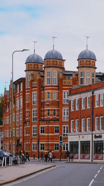 A street view in Harringay, N4, showing a row of narrow, multi-storey terraced houses constructed from red brick with some white and grey accents, alongside modern buildings. The pavement is bustling 