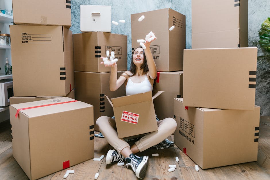 A man and a woman are carrying cardboard boxes down a wooden staircase inside a residential property, preparing for a home relocation or moving process. The man, wearing a dark T-shirt and beige trous