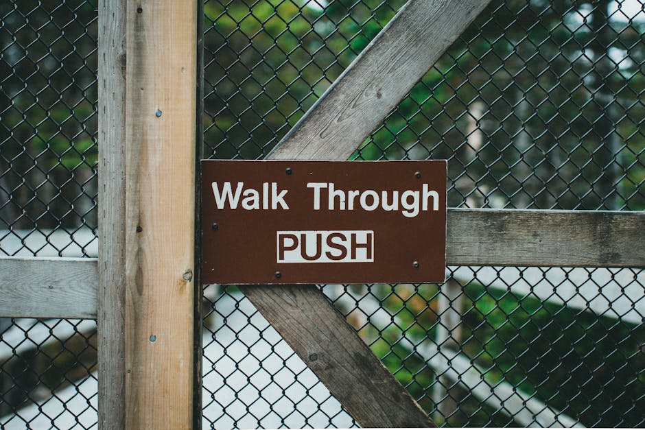 Close-up of a brown metal sign attached to a wooden fence panel, displaying the message 'Walk Through PUSH' in white text. The fence is constructed from horizontal and vertical wooden slats, with a chain-link fence behind it. The background features blurred green foliage and snow-covered ground, indicating an outdoor setting. The sign instructs visitors or movers to push the gate or door to gain access, which is relevant to home relocation and furniture transport processes handled by Man and a Van Haringey, as part of their removals services. The lighting is natural, and the sign is positioned at the intersection of the wooden fence and chain-link fencing, emphasizing the need for proper handling during loading or entry procedures into property grounds.