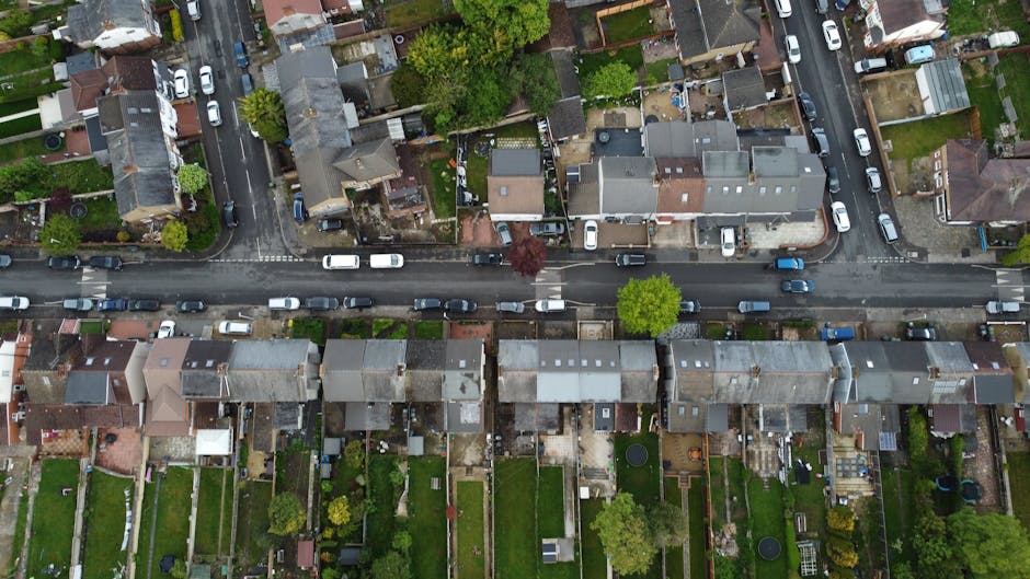 An aerial view of a residential street showing a row of terraced houses with pitched roofs, parked cars lining both sides of the road, and green gardens behind the houses. The image captures the typical tight access properties in Haringey, with narrow pavements and limited space for large moving vehicles. Several trees and small backyard areas are visible, along with garden furniture and decorative elements. The scene is well-lit, with natural daylight highlighting the rooftops and street layout, illustrating the logistical challenges of home relocation and furniture transport in tightly spaced urban environments. This setting exemplifies the kind of residential area where Man and a Van Haringey provides house removal and packing services, managing small streets and confined access points for efficient moving processes.